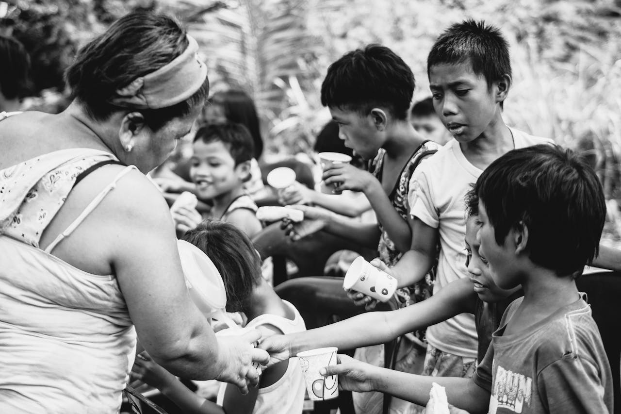 A woman hands out drinks to a group of children outdoors, illustrating community support.